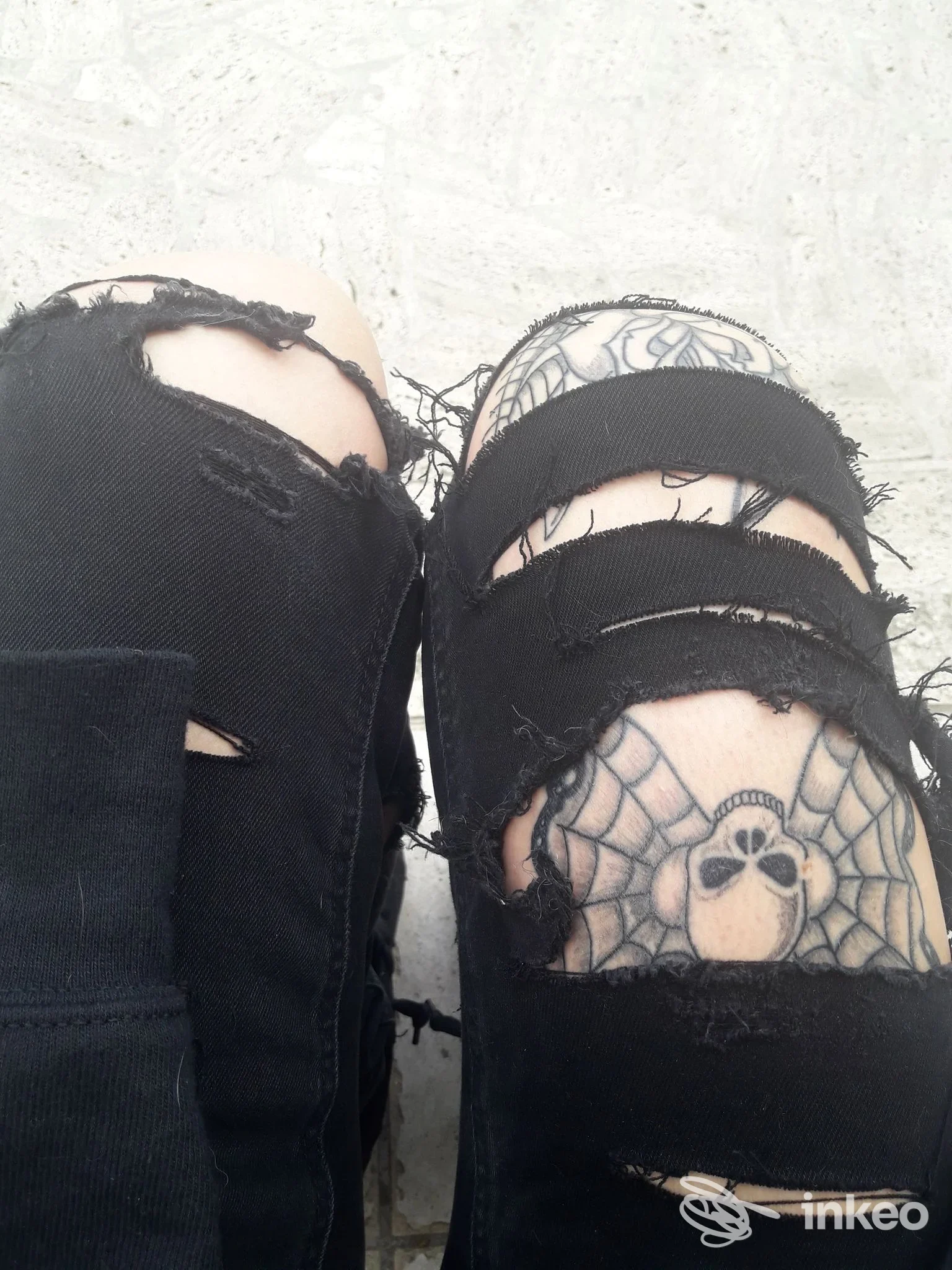 Due ginocchia con jeans strappati che mostrano una tattoo sul ginocchio, ritratto in stile spider web con personaggio centrale, su pelle chiara.