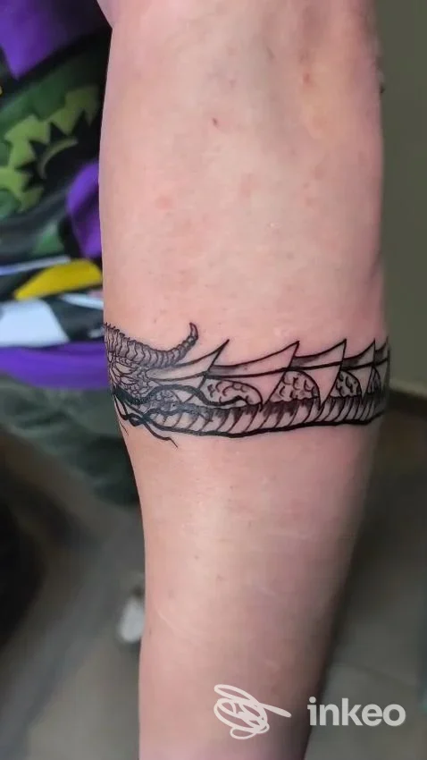 Particolare di un tatuaggio a fascia nera con motivi a squame attorno al braccio, visibile su pelle chiara.