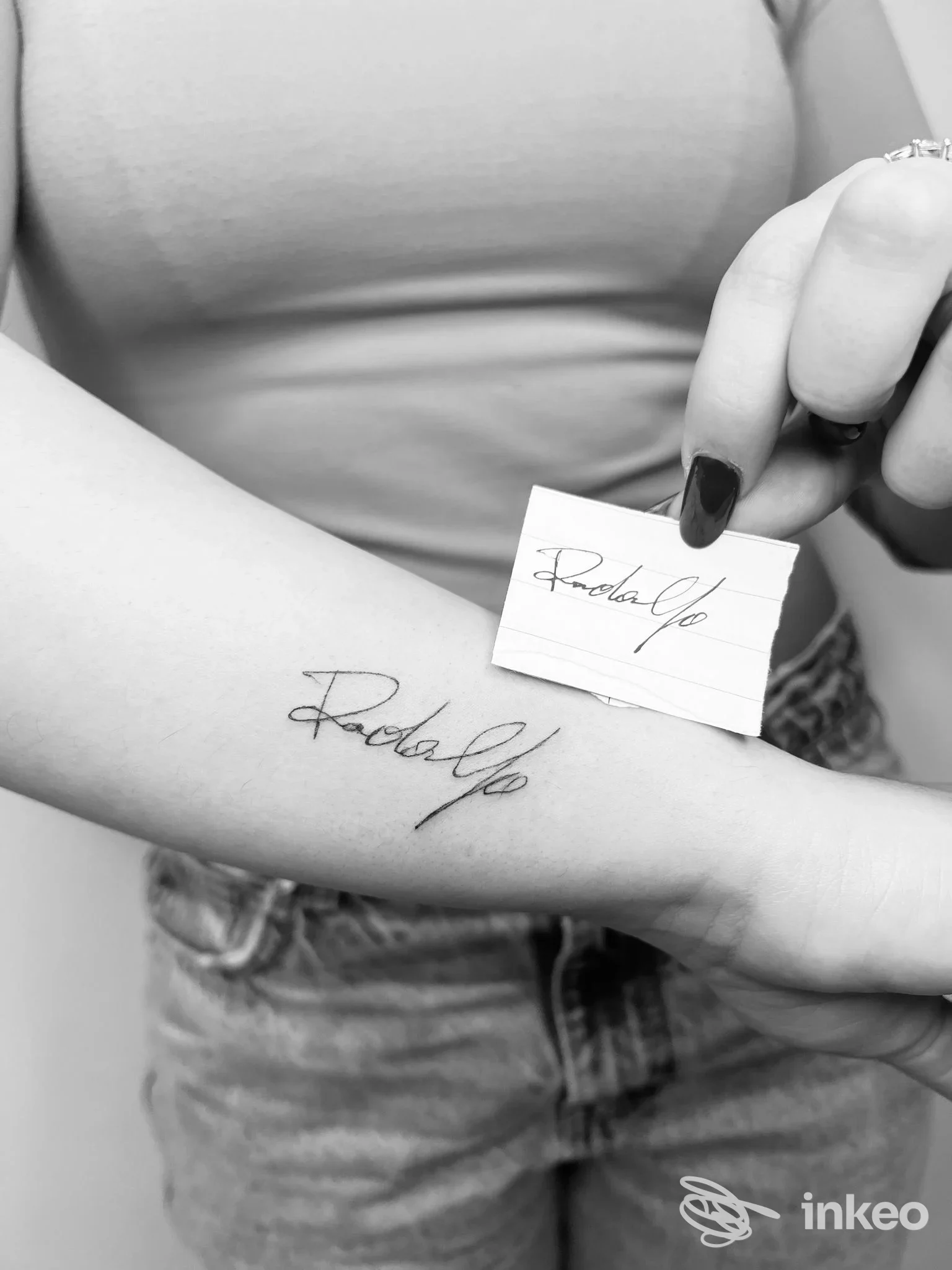 Braccia di una persona con un tatuaggio in corsivo sulla pelle e una piccola carta con firma tenuta vicino al tatuaggio, in bianco e nero, descrivibile come mostrazione della dermopigmentazione realizzata.