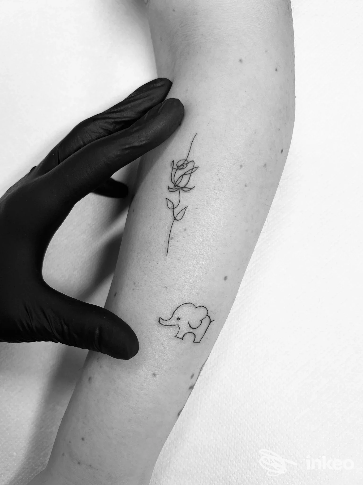 Due braccia visibili: una mano con guanto nero sfiora l'avambraccio rivelando due tatuaggi: una rosa stilizzata in alto e un piccolo elefante in basso, sfondo bianco.