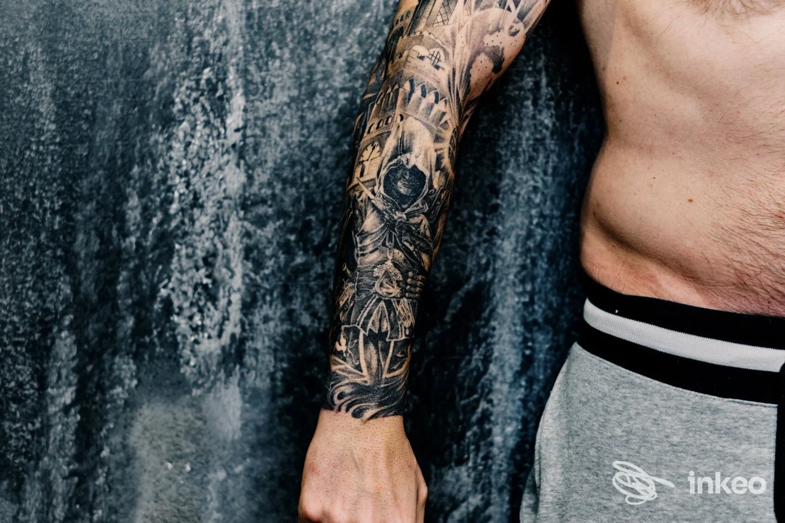 Avambraccio sinistro con tatto pieno di stile tribale e motivi decorativi, visibile su sfondo grigio, torso nudo parziale.