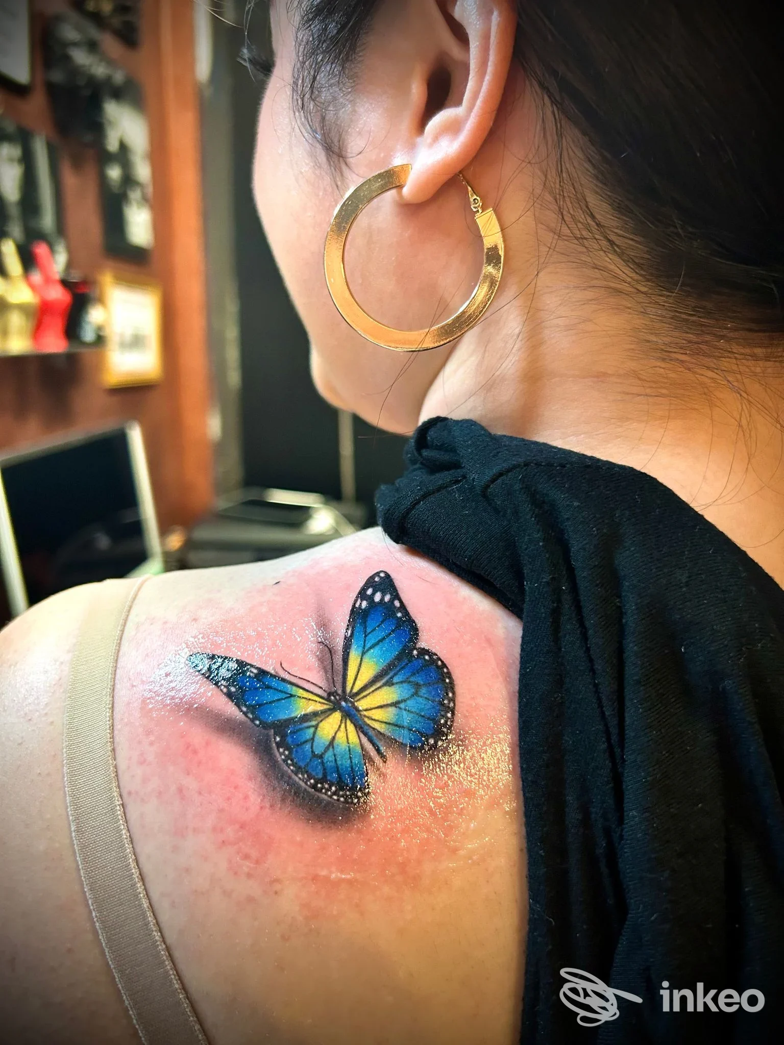 Tattoo reale di una farfalla blu e gialla sulla spalla, contorno pelle arrossata, visibile parte del volto sullo sfondo.