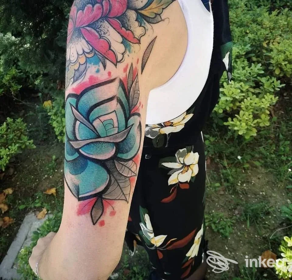 Braccio di una persona con un grande tatuaggio colorato a tema floreale, visibile su sfondo esterno con abbigliamento estivo, firma di mano manos Hand Made Tattoo Shop, Avezzano.