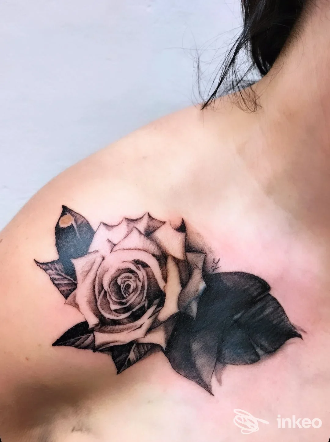Tattoo realistico di rosa e foglie nere-grigie su spalla, realizzato sulla pelle, senza volto visibile.