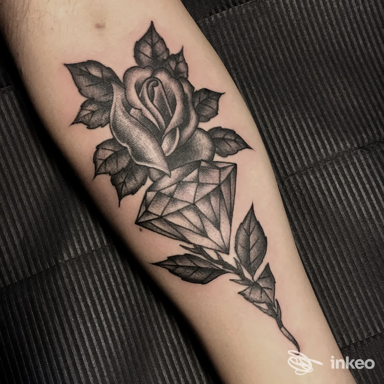 Tatuaggio realistico in bianco e nero sull’avambraccio: rosa con foglie e un parallelepipedo the diamond, realizzato sulla pelle.
