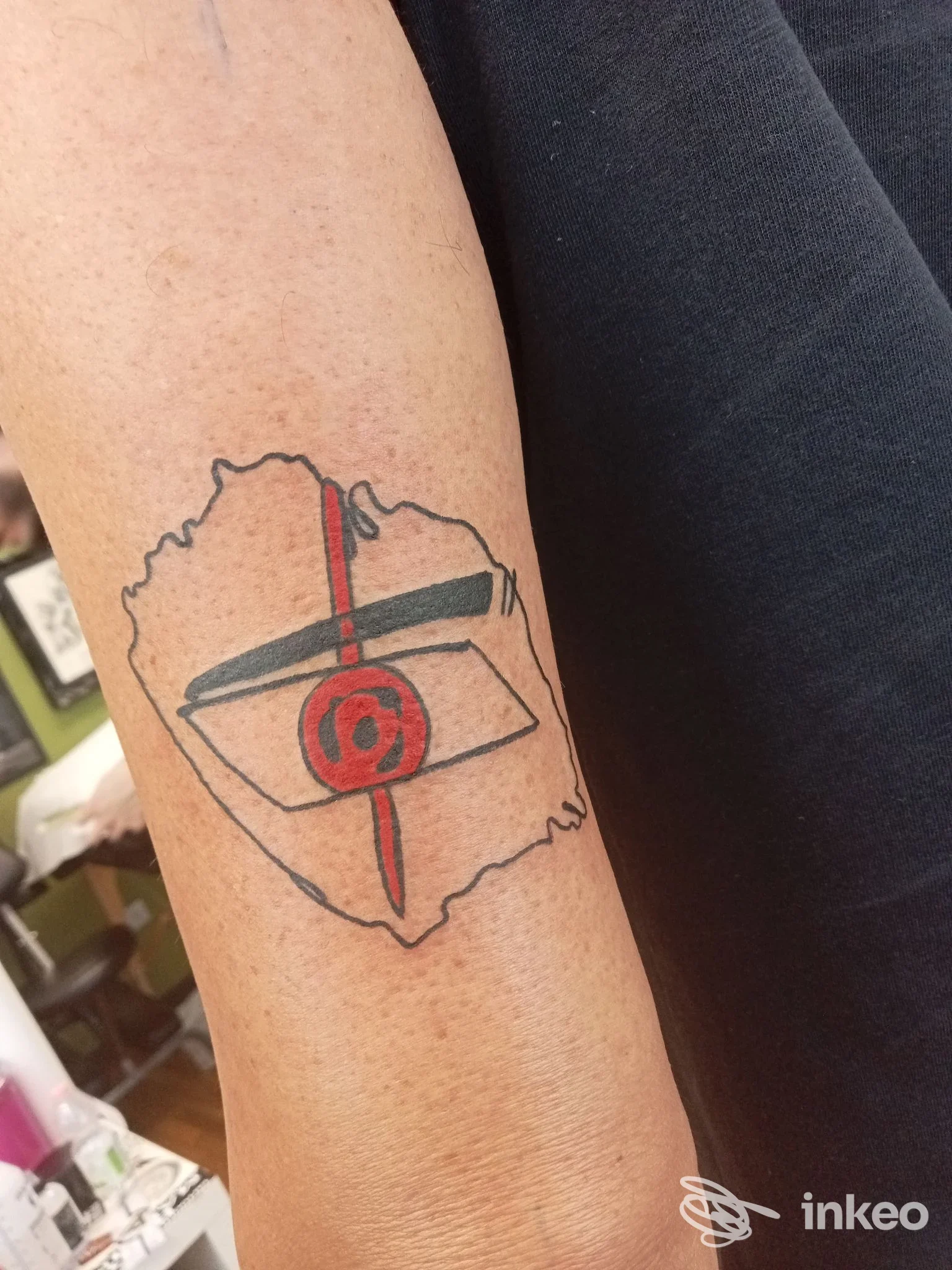 Tatuaggio visibile sull'avambraccio raffigurante una scatola con simbolo rosso e linee nere, contornata da una forma astratta, realizzato sulla pelle della persona.