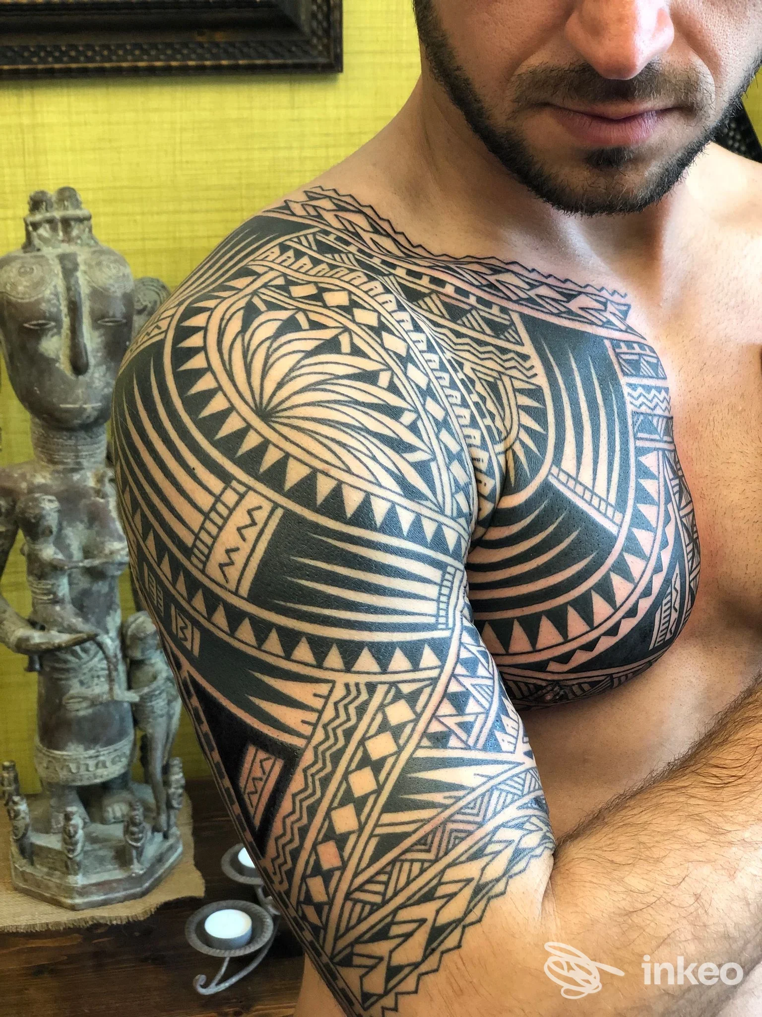 Braccio e spalla destra visibili di un uomo, tatuaggio tribale geometrico nero molto articolato che copre braccio, spalla e petto superiore, con sfondo interno giallo e oggetti decorativi.