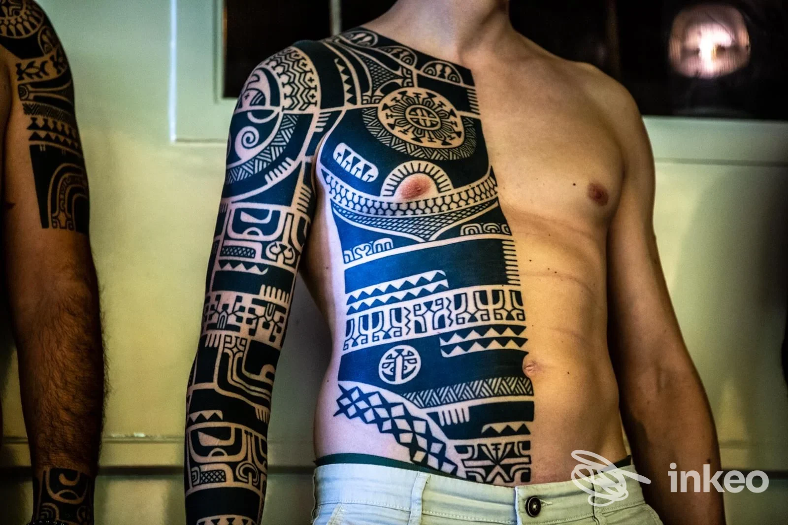 Primo piano di un uomo senza viso visibile in foto, mostra un grande tatuaggio nero tribale che copre braccio e torso sinistro, scollo scoperto e pantaloni chiari, studio Wallace Tattoo a Milano.