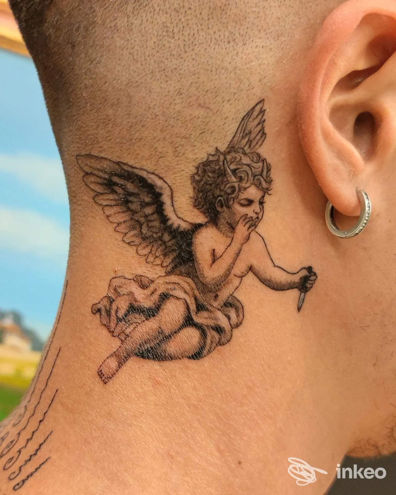 Tattoo corporale raffigurante un cherubino alato sul collo, visibile su pelle reale, con orecchino visibile sull'orecchio destro; nessun overlay o immagine esterna.