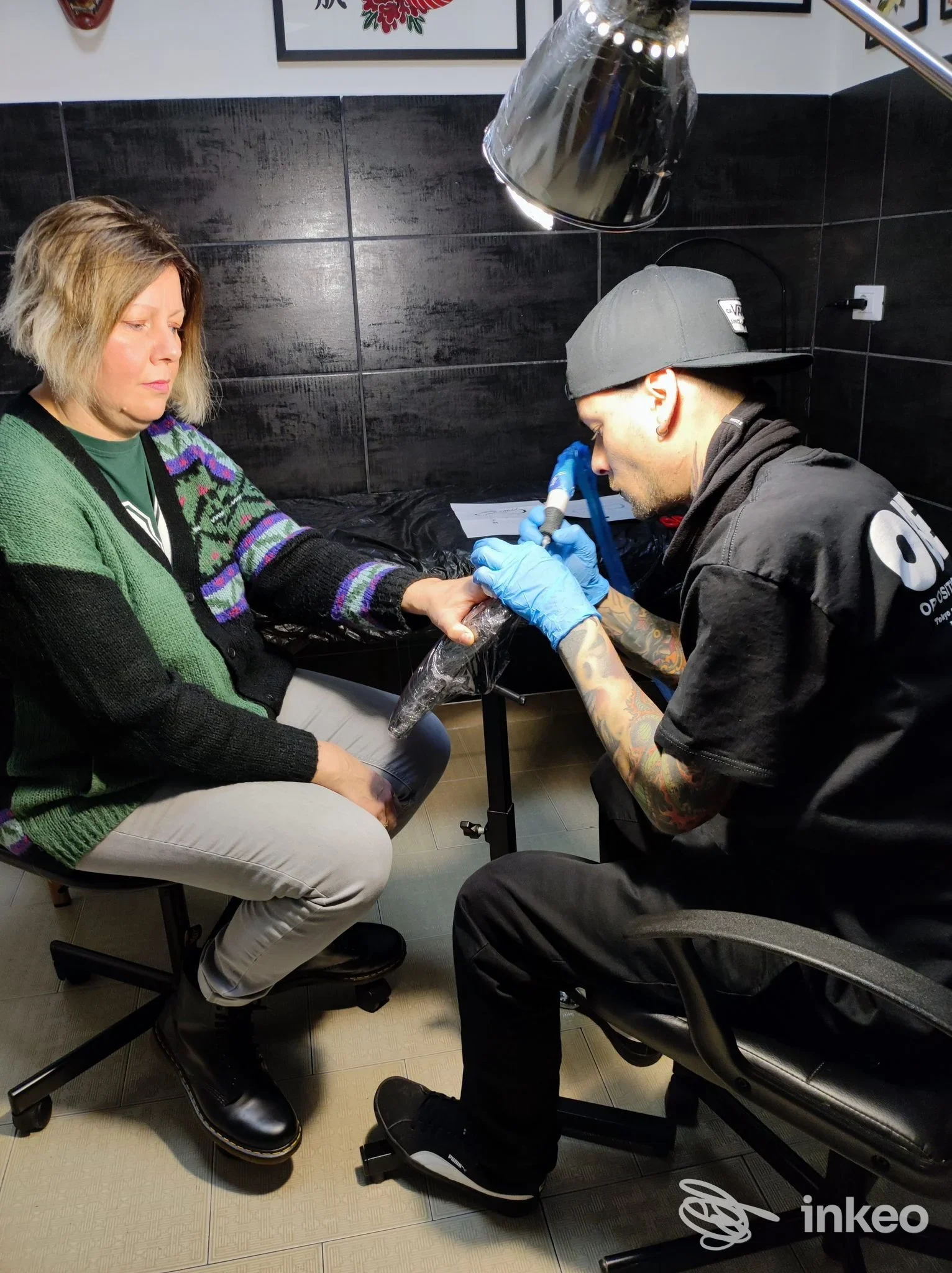 Tattoo in corso su una persona seduta, mano dell’operatore tatuatore con guanti blu lavora sull’avambraccio; studio ink e pareti scure sullo sfondo, illuminazione da lampada al neon.