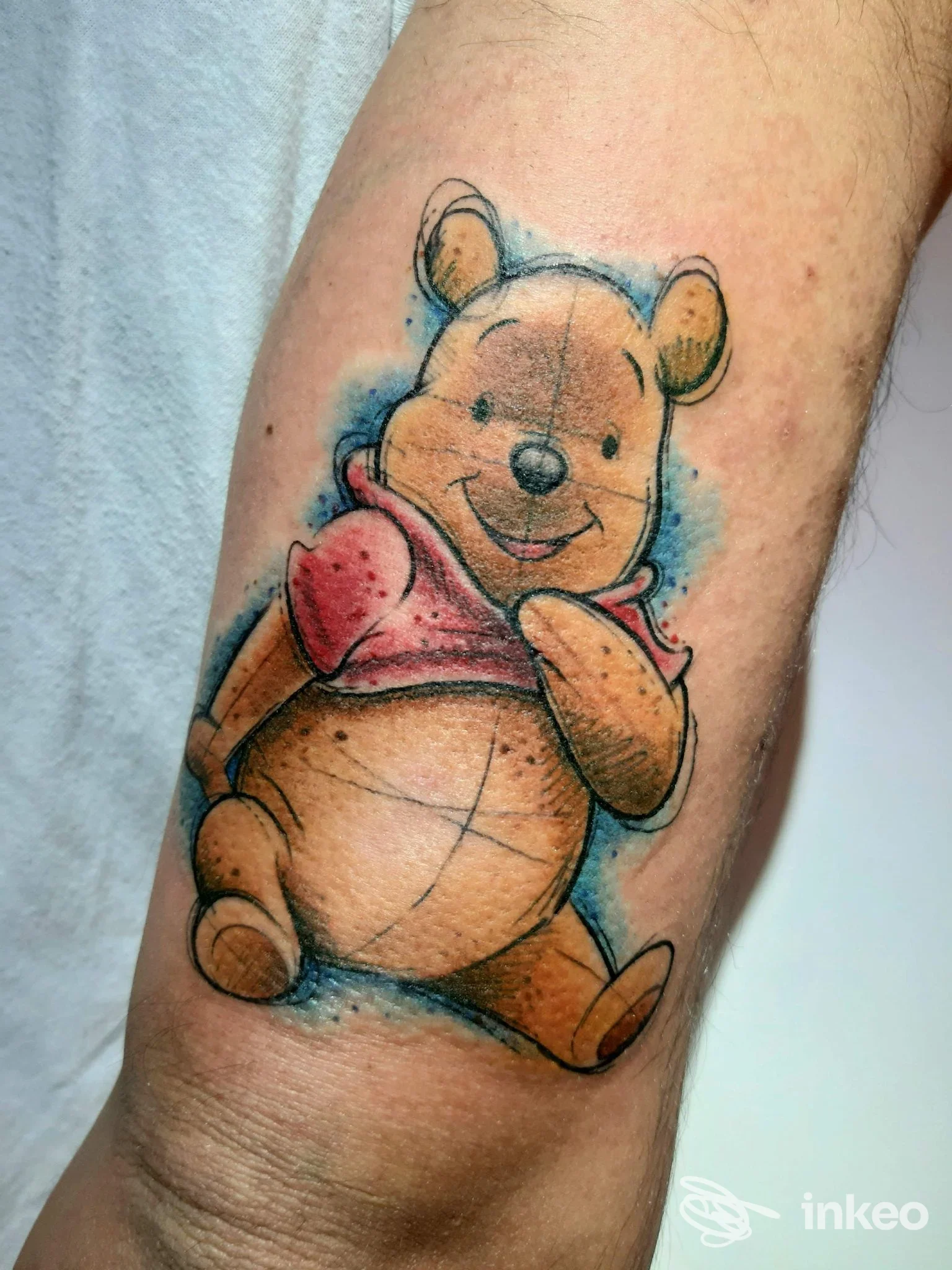 Tattoo a colori di Winnie the Pooh sull’avambraccio, stile cartone animato, con sfondo blu. braccio visibile, nessun overlay social, composizione chiara.