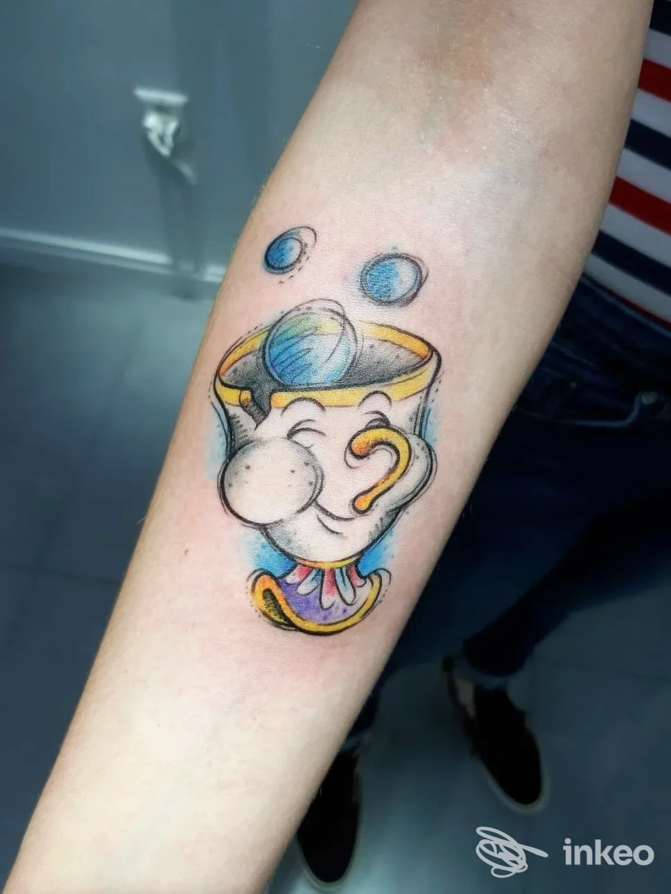 Tatuaggio su avambraccio raffigurante una tazza con cappello e personaggio colorato, realizzato presso New Moon Tattoo Studio a Chieti.