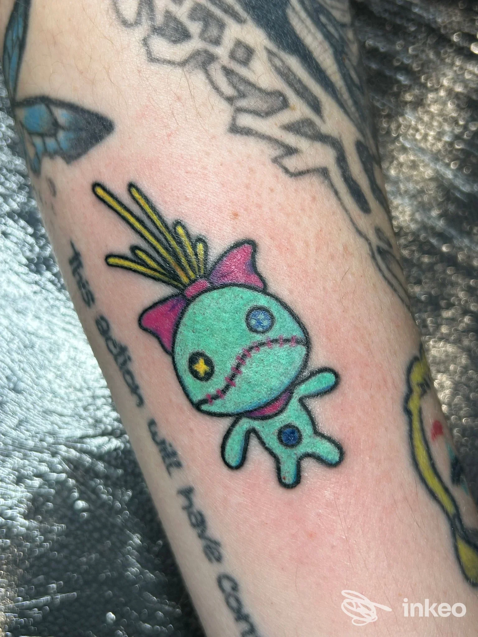 tatuaggio colorato su pelle che raffigura un personaggio cartone animato verde con occhi colorati e dettagli rosa, su braccio/avambraccio; stile fumettoso.