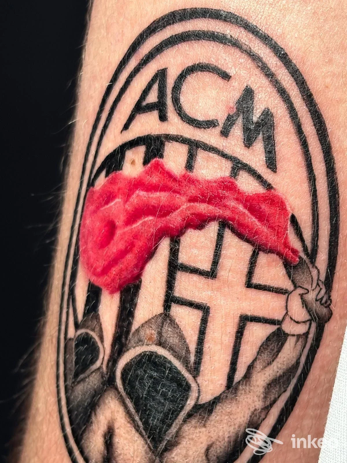 Tatuaggio visibile su pelle, con lettere ACM all'interno di una cornice ovale, con segni rossi sopra la linea centrale. studio Splat Ink TattooShop, Vimercate.