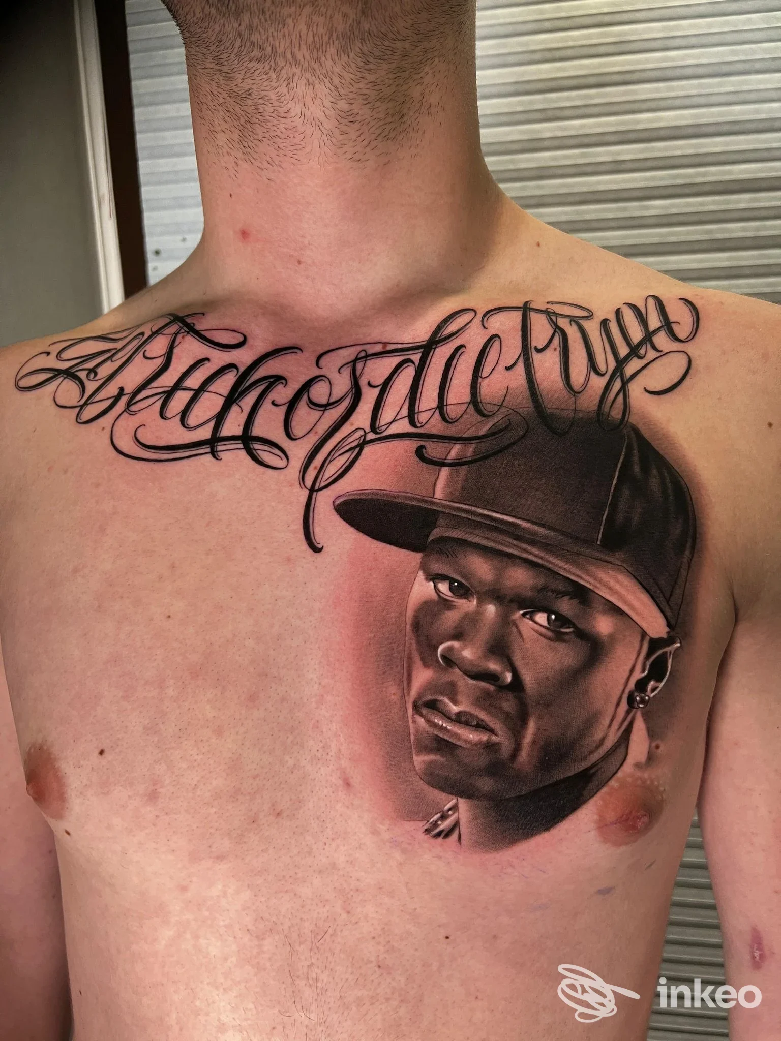 Tattoo a colori sul torace raffigurante un volto maschile con cappello, abbinato a scritte elaborate sulla clavicola destra; la pelle è visibile e il tatuaggio copre la zona pettorale.