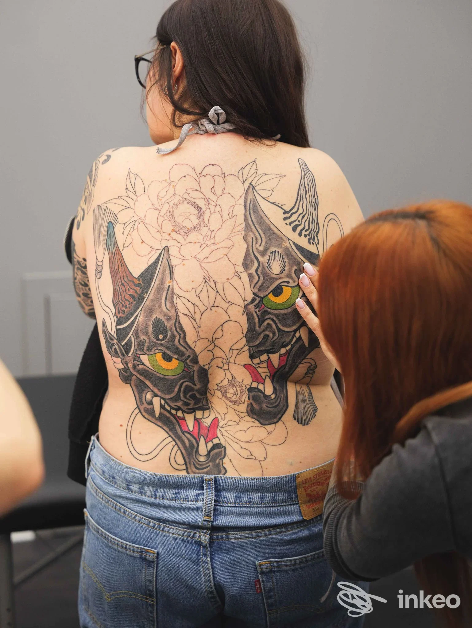 Schiena di una persona con grande tatuaggio realistico di due gatti con occhi gialli, ornamenti floreali, supportato da un secondo tatuaggio sul fianco. la scena mostra una correzione di un collega sul tessuto cutaneo.