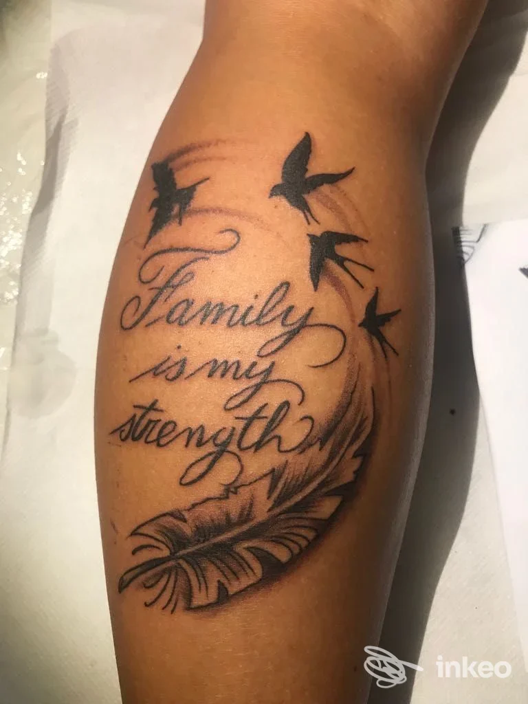 Tattoo sul braccio con testo scritto in corsivo "Family is my strength" accompagnato da uccelli neri in volo e una piuma decorativa.