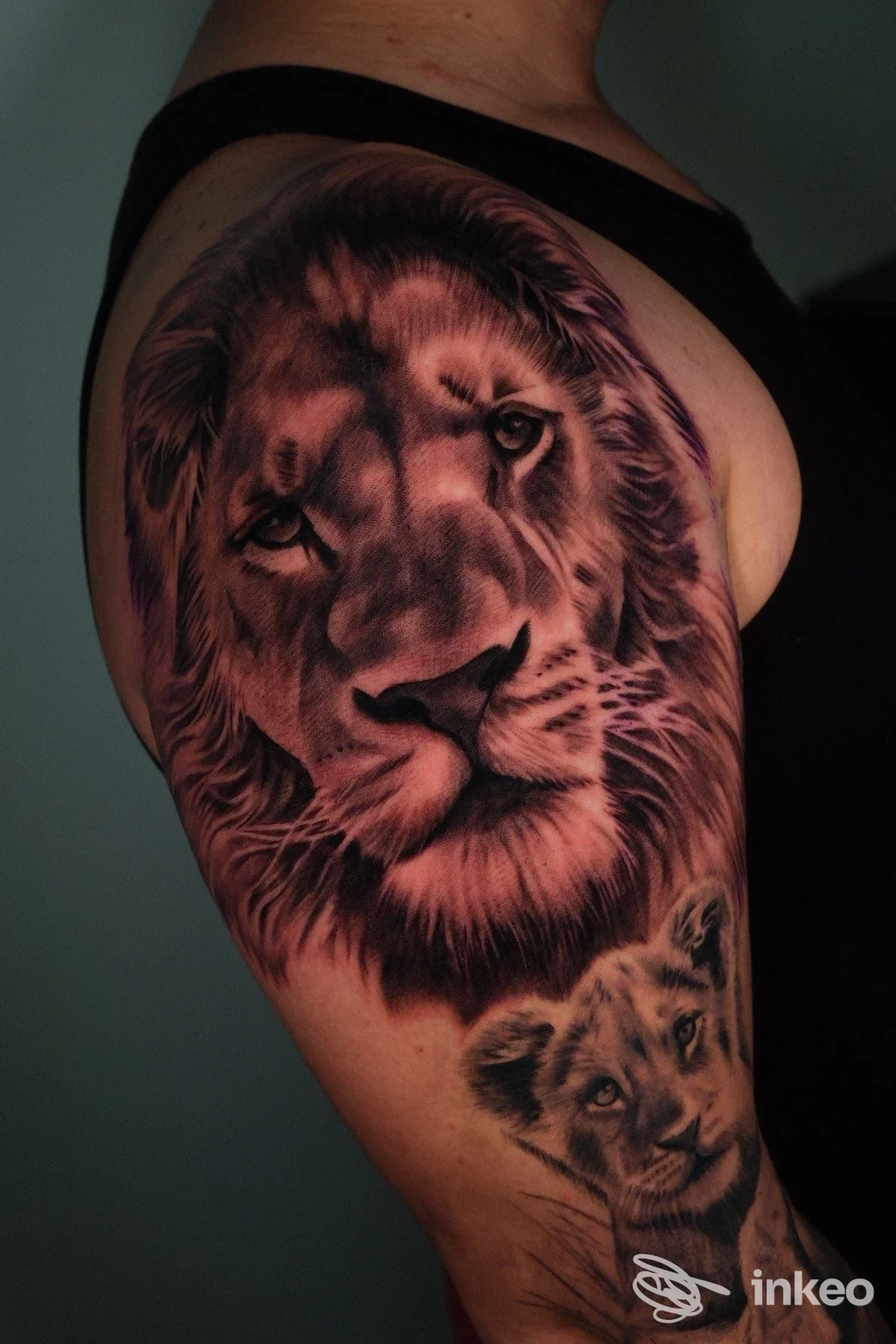 Tattoo realistico di leone e cucciolo sul braccio, tonalità nero-grigio, visibile pelle e ombreggiatura dettagliata.
