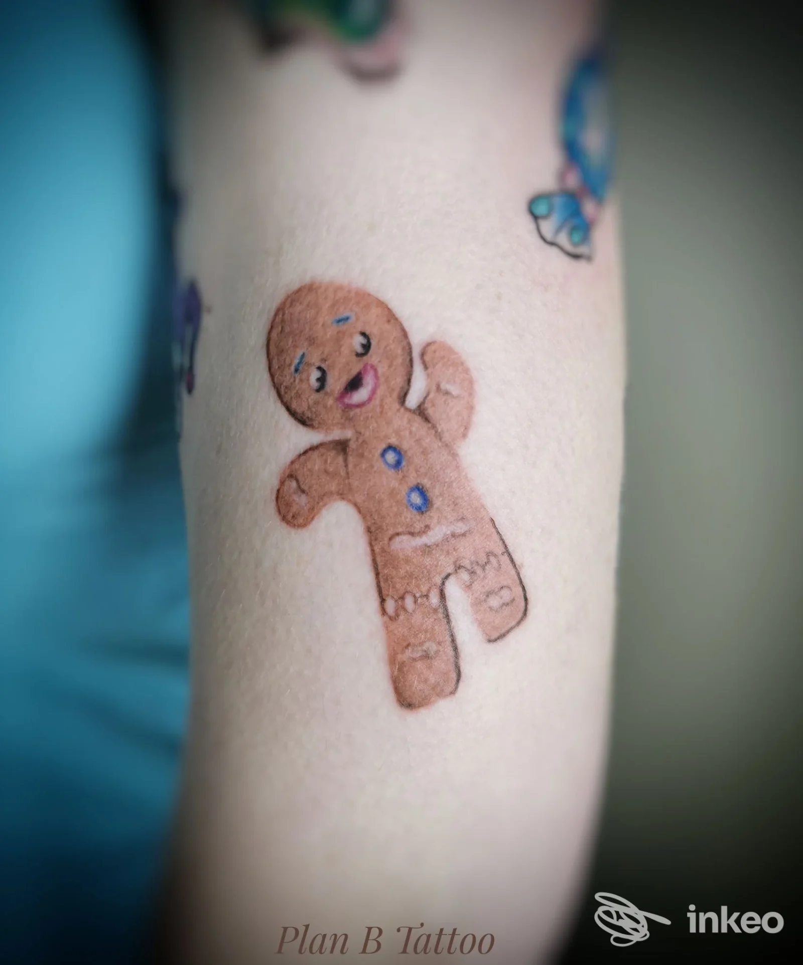 Tattoo su pelle di un braccio: omino di pan di zenzero marrone con sorriso, due bottoni blu, accompagnato da altri disegni colorati visibili ai margini.