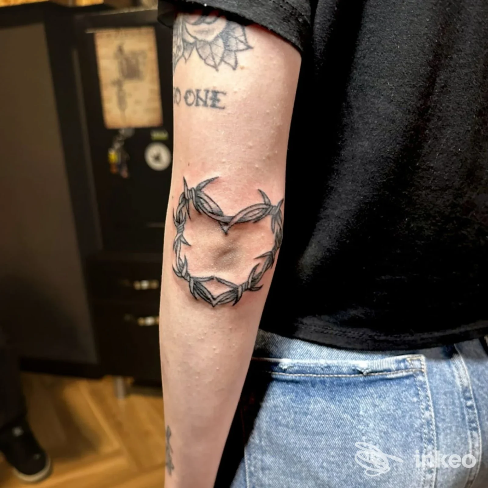 Dettaglio di un braccio: tatuaggio a forma di cuore realizzato con filo spinato sull'avambraccio interno sinistro, tonalità nere e grigie, tessuto nero della maglietta visibile.
