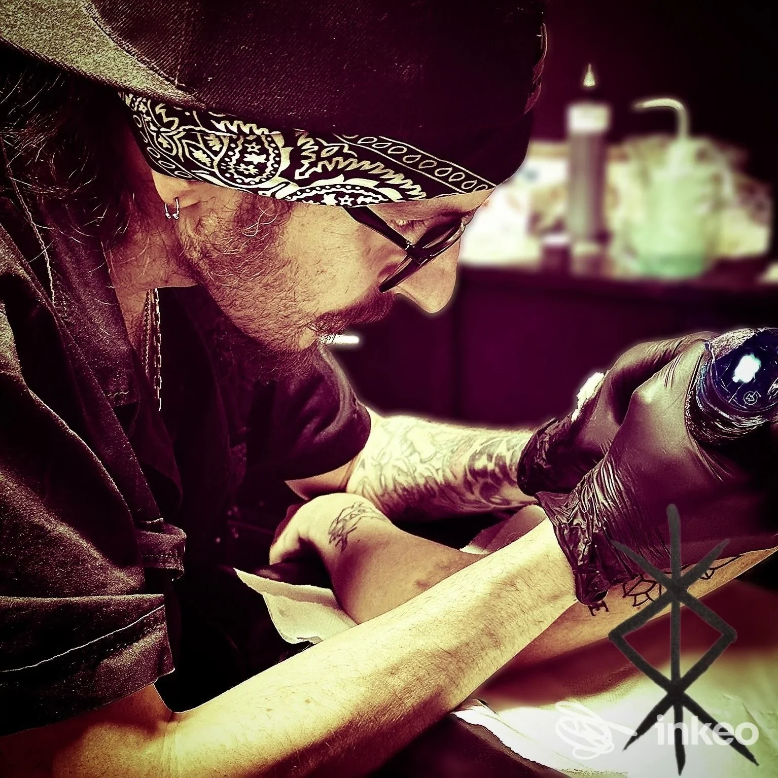 Un tatuatore con bandana sta tatuando il braccio di un cliente in uno studio, con strumenti visibili e attrezzature sul tavolo sullo sfondo, in Atessa, Memento Tattoo Parlor.