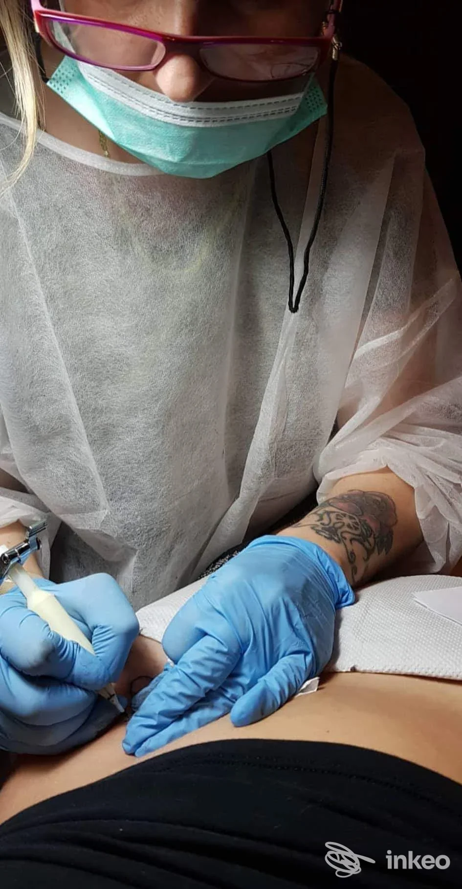 Una professionista con mascherina e guanti blu esegue una tatuatura su pelle nuda, con strumenti in mano, in abito protettivo trasparente.