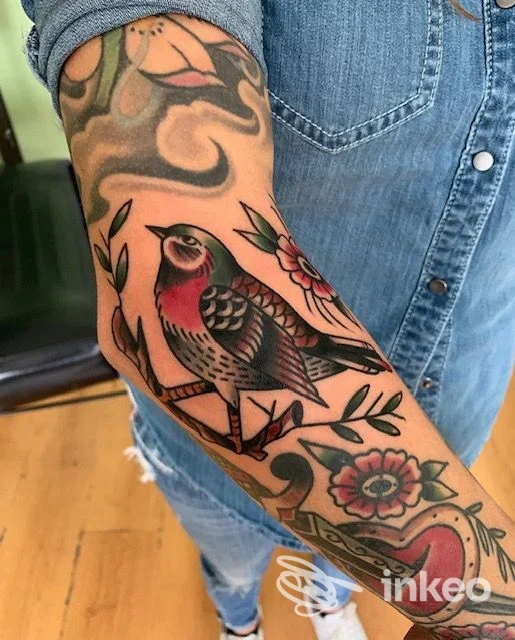 Braccio di una persona con tatuaggio completo: un uccello stilizzato al centro, fiori e foglie, con sfondo di jeans; colori vividi e linee nette.