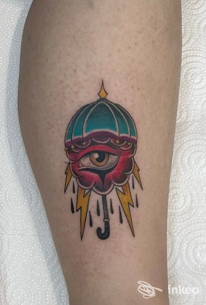Tatuaggio su avambraccio: un ombrello color teal con al centro un occhio rosso, circondato da elementi gialli e una goccia nera in basso.