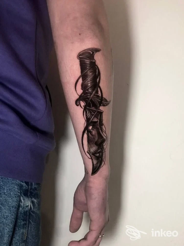 Avambraccio di una persona con un disegno di tatuaggio nero a forma di lama o lama intrecciata, realizzato sulla pelle, sfondo neutro.