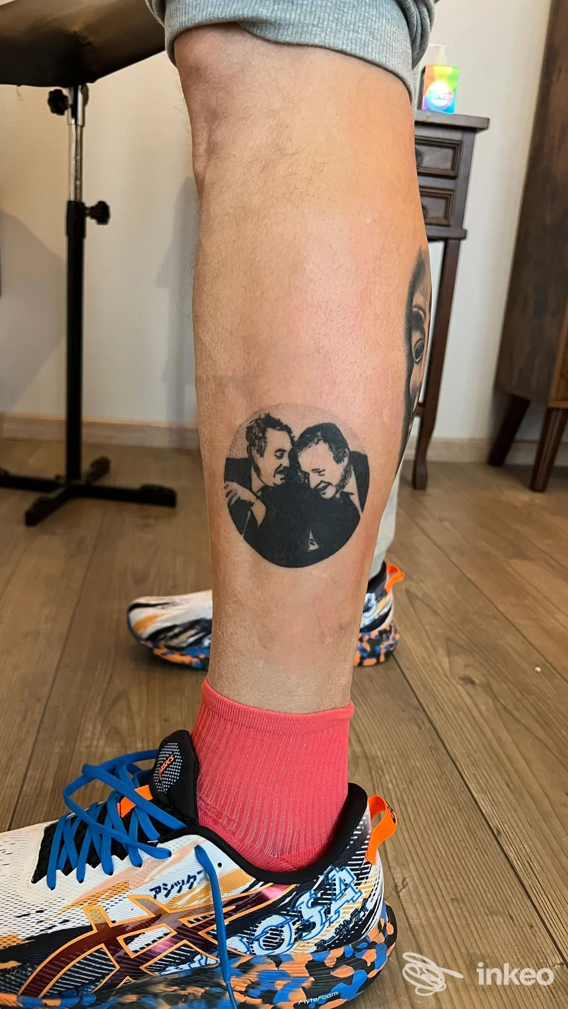 Tatuaggio circolare sul polpaccio visibile: due persone in una scena in bianco e nero all'interno di un cerchio, su gamba nuda vicino al piede con sneaker colorate e calzini rosa.