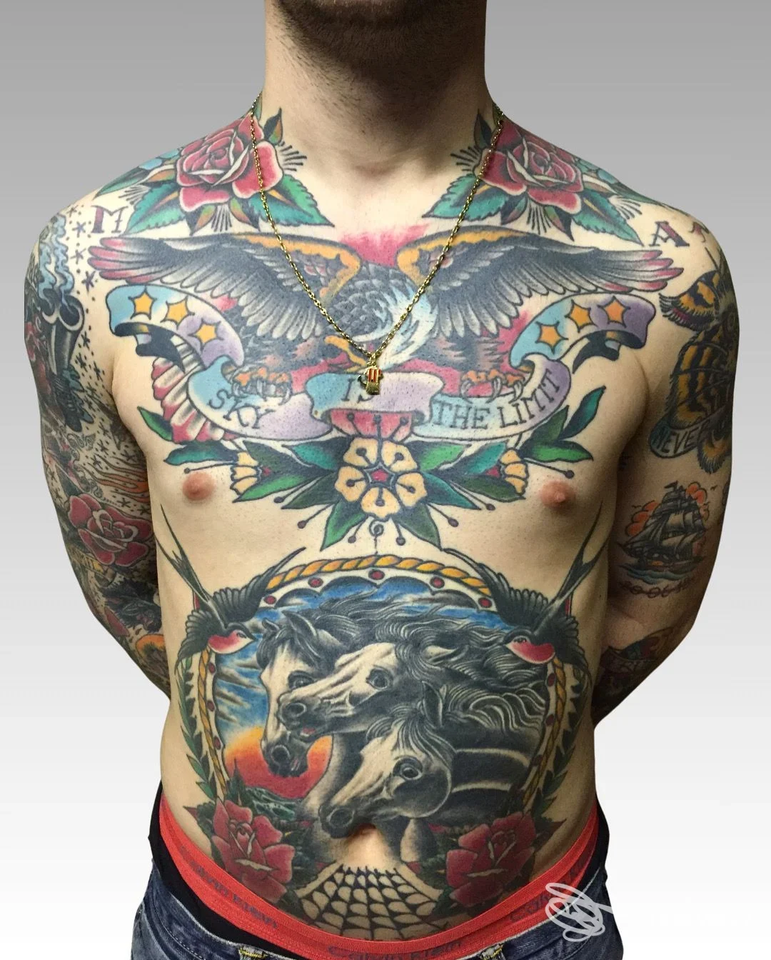 Torso nudo di un uomo coperto da tatuaggi colorati: pettorale, addome e braccia pieni di disegni, collana con ciondolo visibile. inizio testa non visibile.
