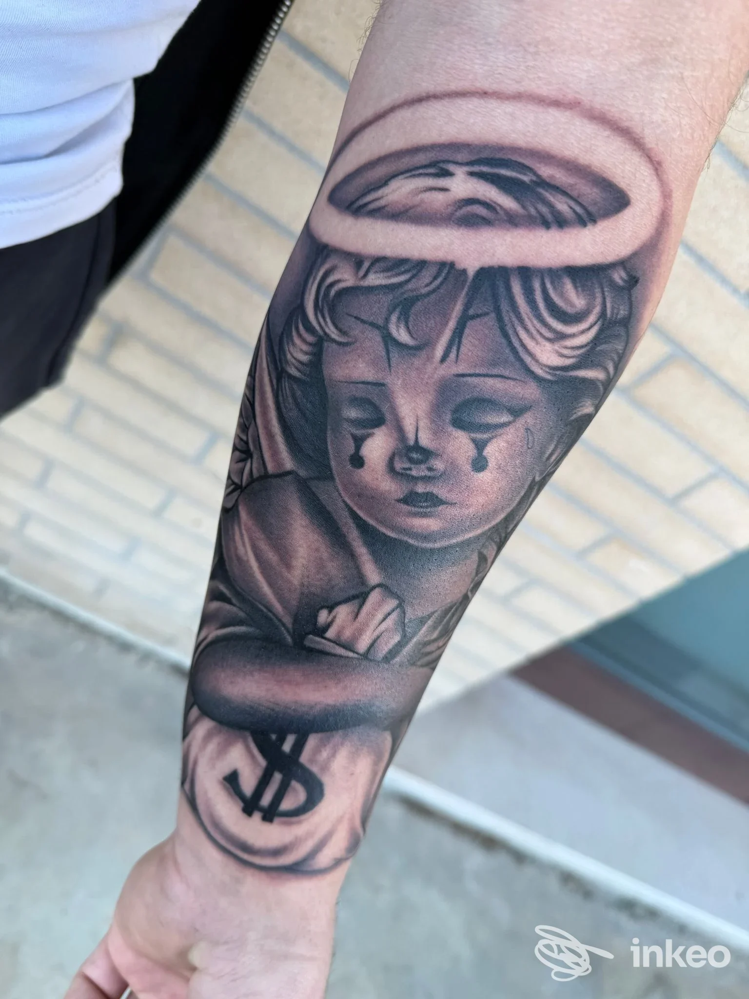 Tattoo su avambraccio raffigurante un angelo con aureola, stile inchiostrato scuro e dettagli realistici, visibile la pelle e il contorno del braccio.