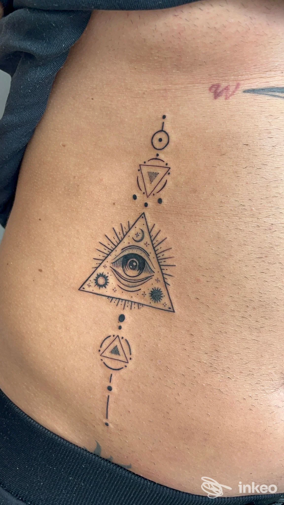 Tattoo geometrico sul fianco: triangolo con occhio centrale, decorazioni e punti lungo una linea verticale, realizzato in stile linea nera su pelle chiara.