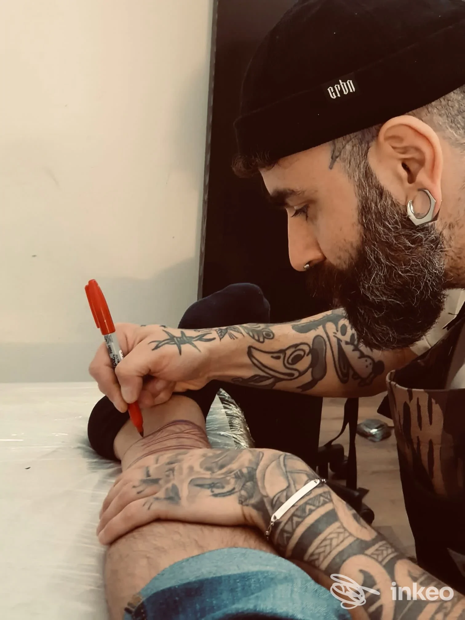 Un tatuatore con cappello e orecchini tatua l’avambraccio di un cliente al Archetipo Tattoo Studio a Teramo, durante la sessione, con linee visibili e strumenti da tatuaggio.