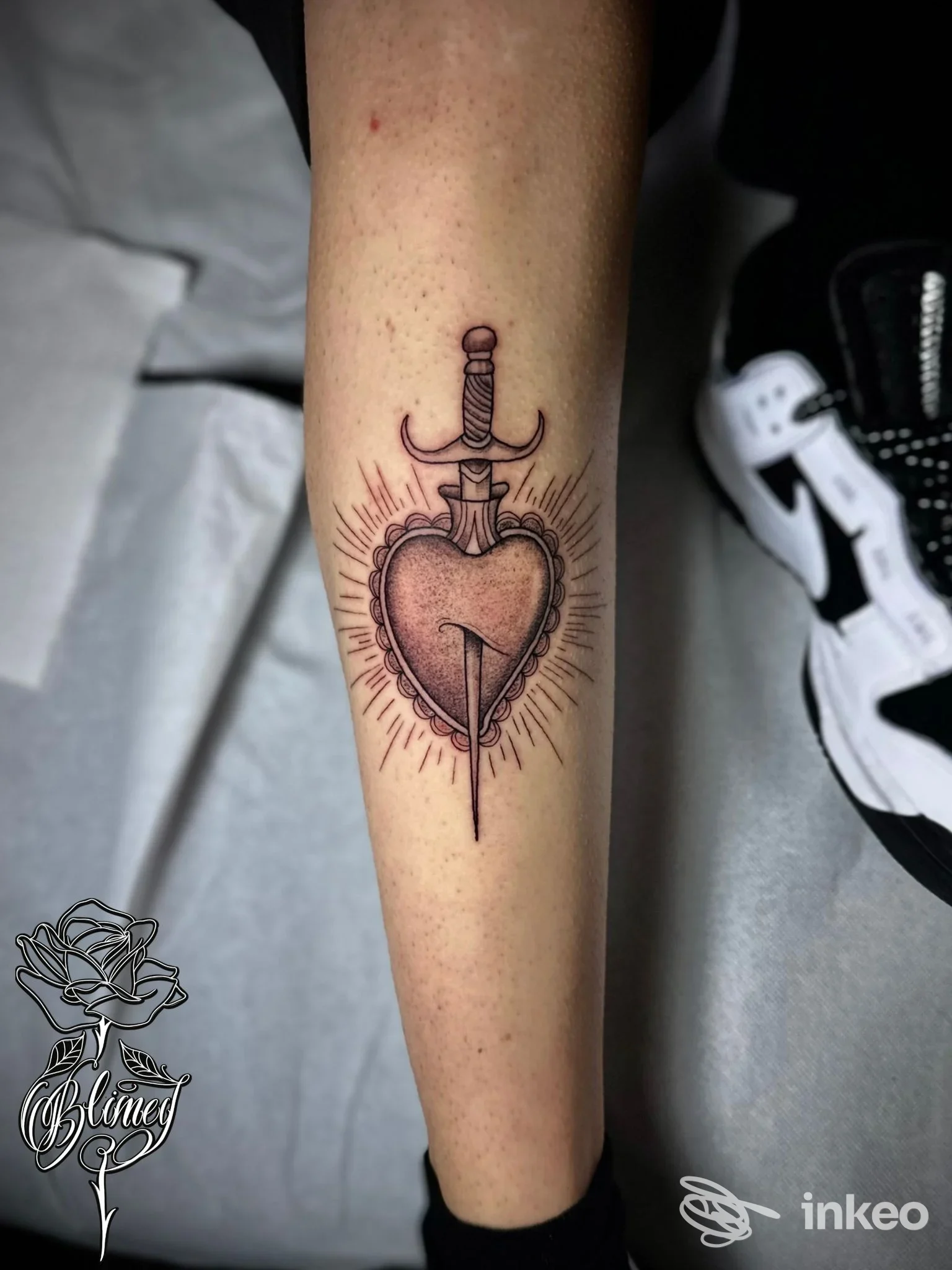 Tatuaggio di cuore trafitto da una spada sul braccio, con radianti intorno al disegno, colori nero e rosso su pelle chiara.