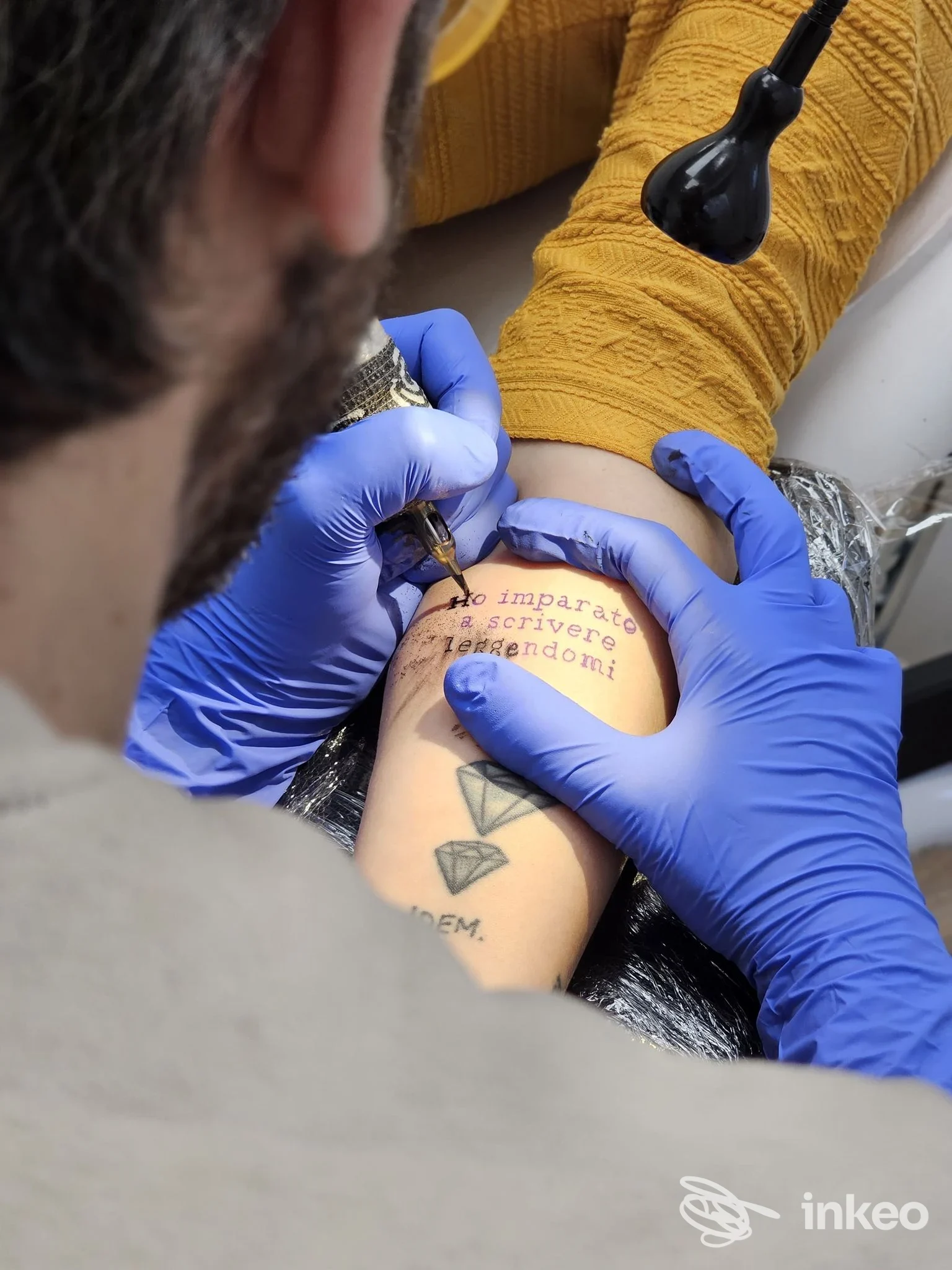 Veduta ravvicinata di una mano guantata che esegue un tatuaggio su un avambraccio, con testo visibile sull’epidermide e una piccola grafica, in studio