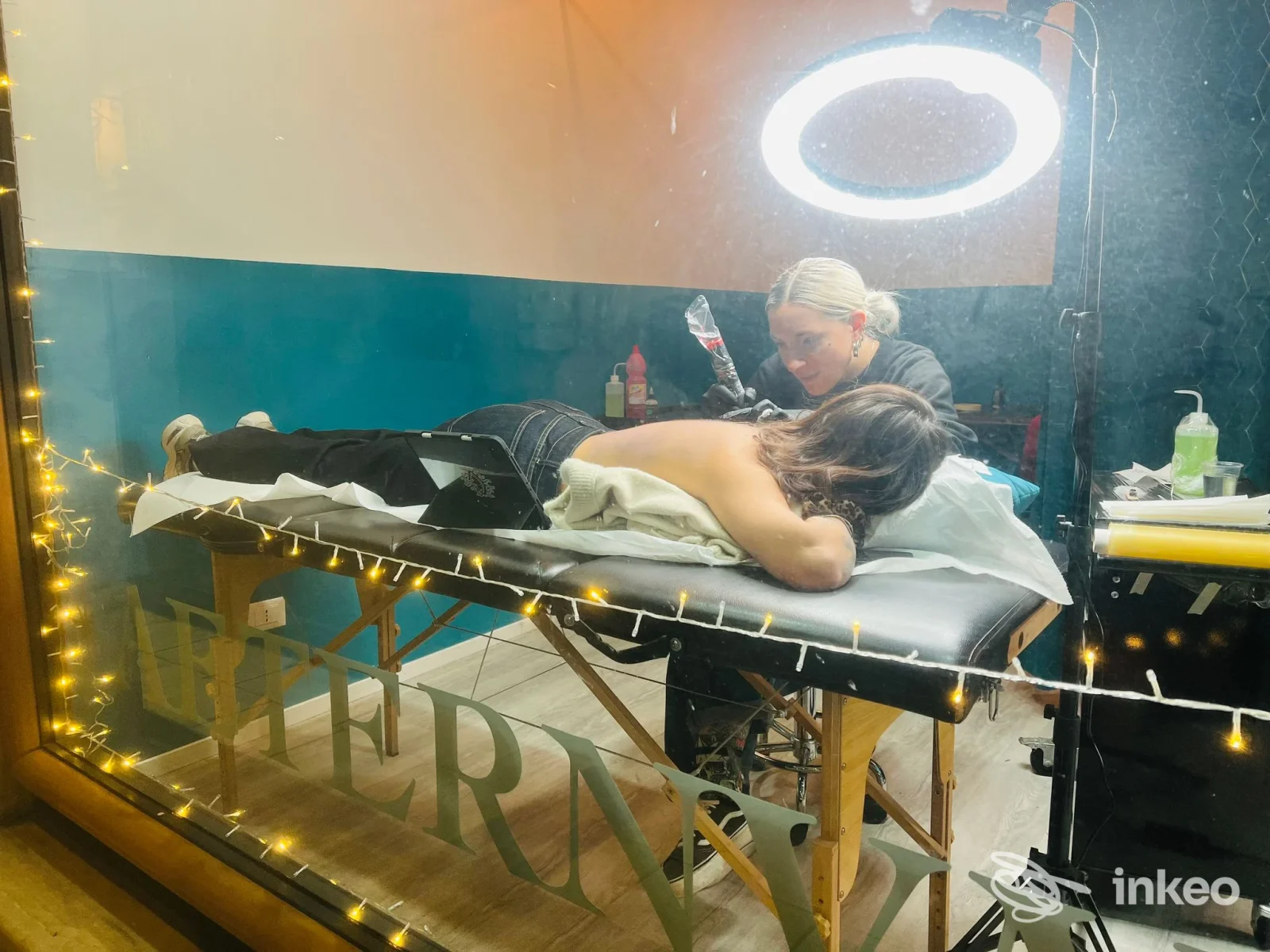 Cliente disteso sul tavolo da tatuaggio nello studio Aeternvm a Pescara, operatore al lavoro con luci circolari e attrezzature visibili.