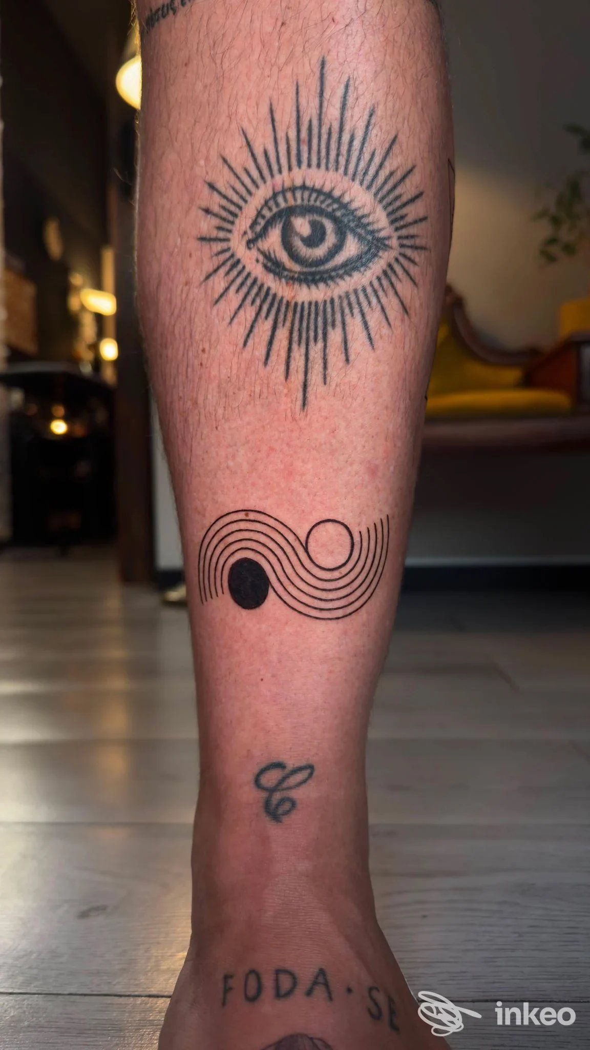Tattoo visibile sulla gamba: un occhio all’interno di un cerchio di raggi e un elemento grafico con linee curve circolari, eseguiti sul polpaccio in pelle nuda.