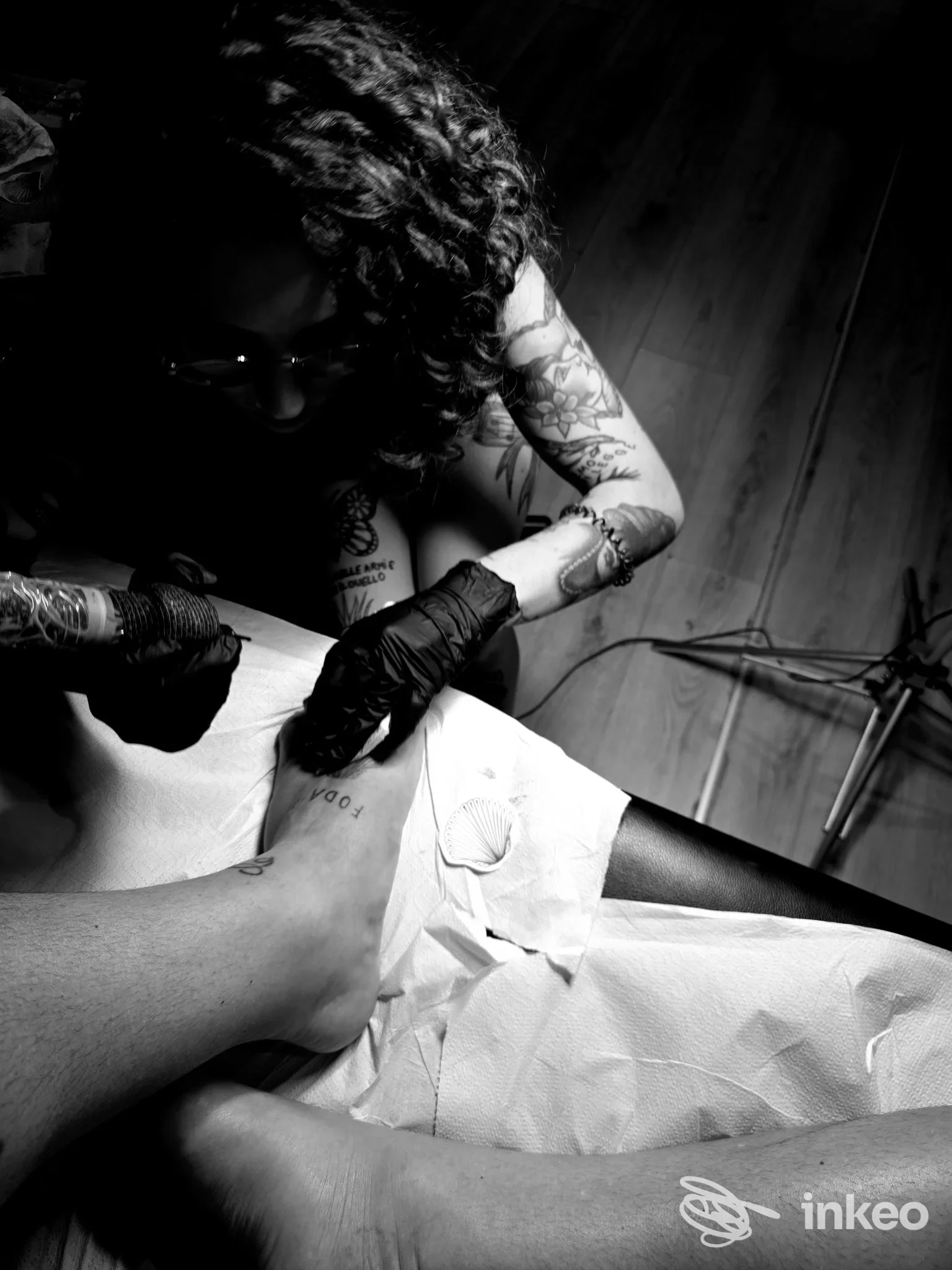In studio Delos a Montesilvano, un tatuatore lavora su una gamba coperta da carta, mentre usa guanti neri e strumenti per tatuaggio in un ambiente professionale in bianco e nero.