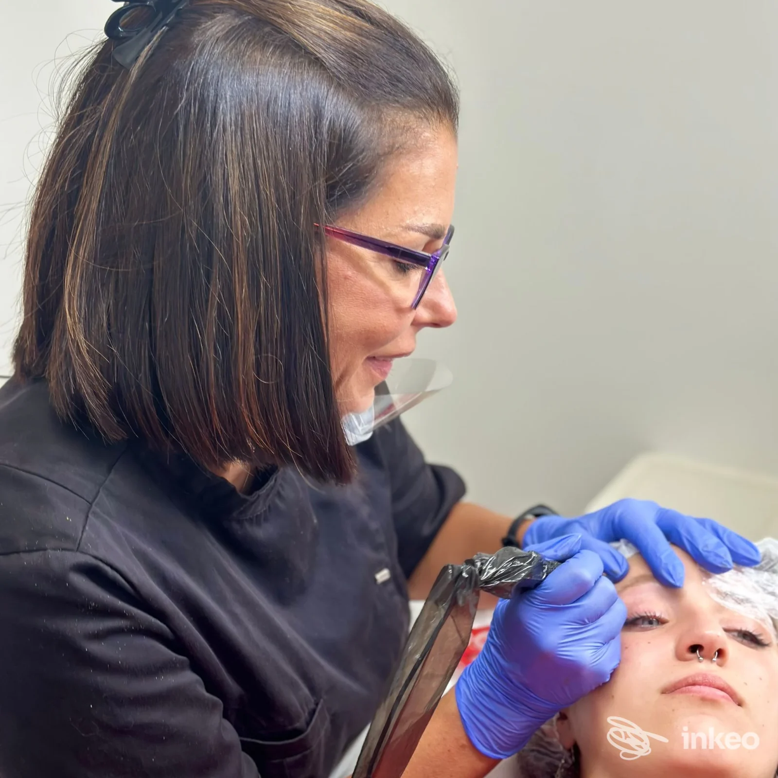 Operatrice con guanti blu esegue trucco permanente su sopracciglio/punto vicino all’occhio di una paziente, seduta su poltrona, in studio Rosalinda Ricci a Pescara.