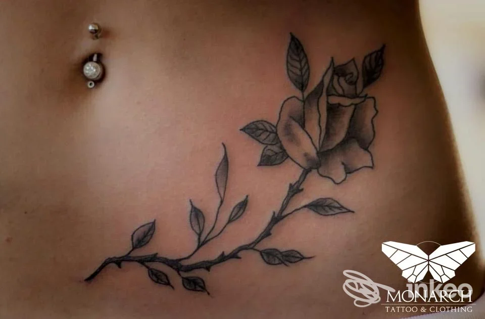 Tattoo reale visibile sulla pelle: ramo di rosa lungo l’addome, con punto di piercing visibile, su pelle chiara, senza volto umano mostrato. studio Monarch Tattoo, Pescara.