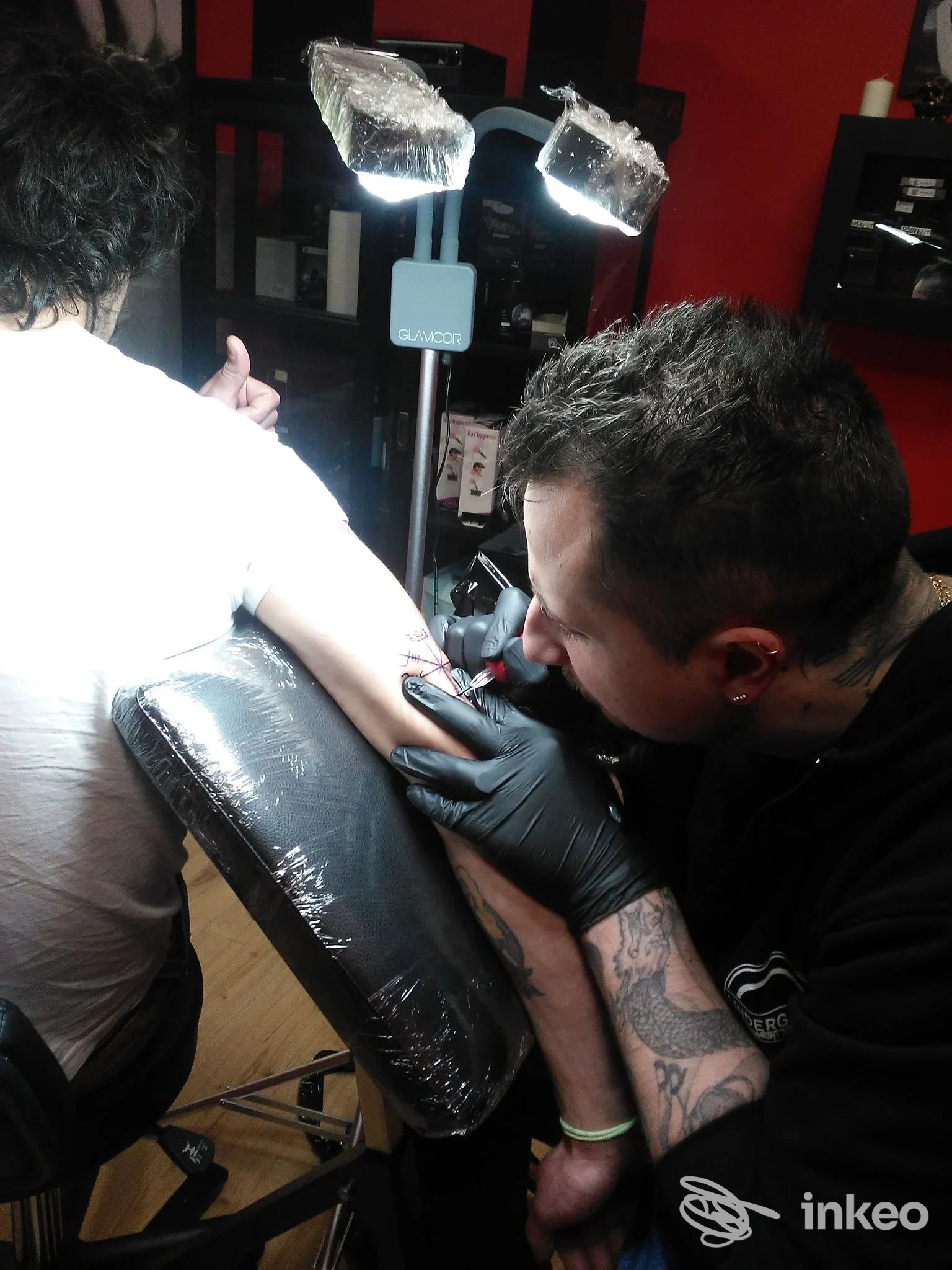 Un tatuatore con guanti neri lavora sul braccio di un cliente all'interno di uno studio, con luci professionali e seduta da tattoo, evidenziata la striscia di pelle tatuata.