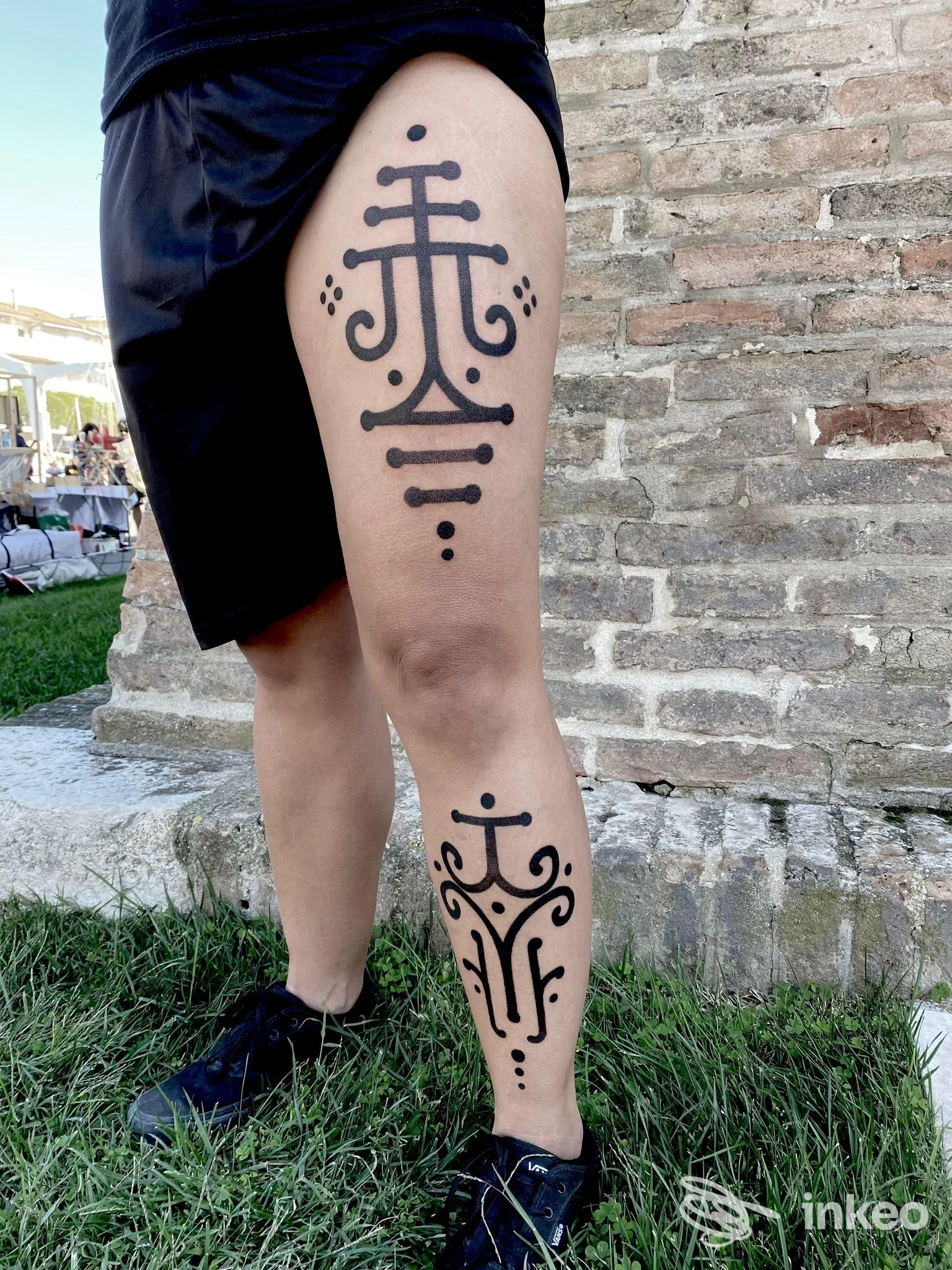 Tattoo elegante sulla gamba sinistra di una persona, con figure lineari nere e motivi circolari, visibile in alto e sotto il polpaccio; sfondo urbano con pietra.