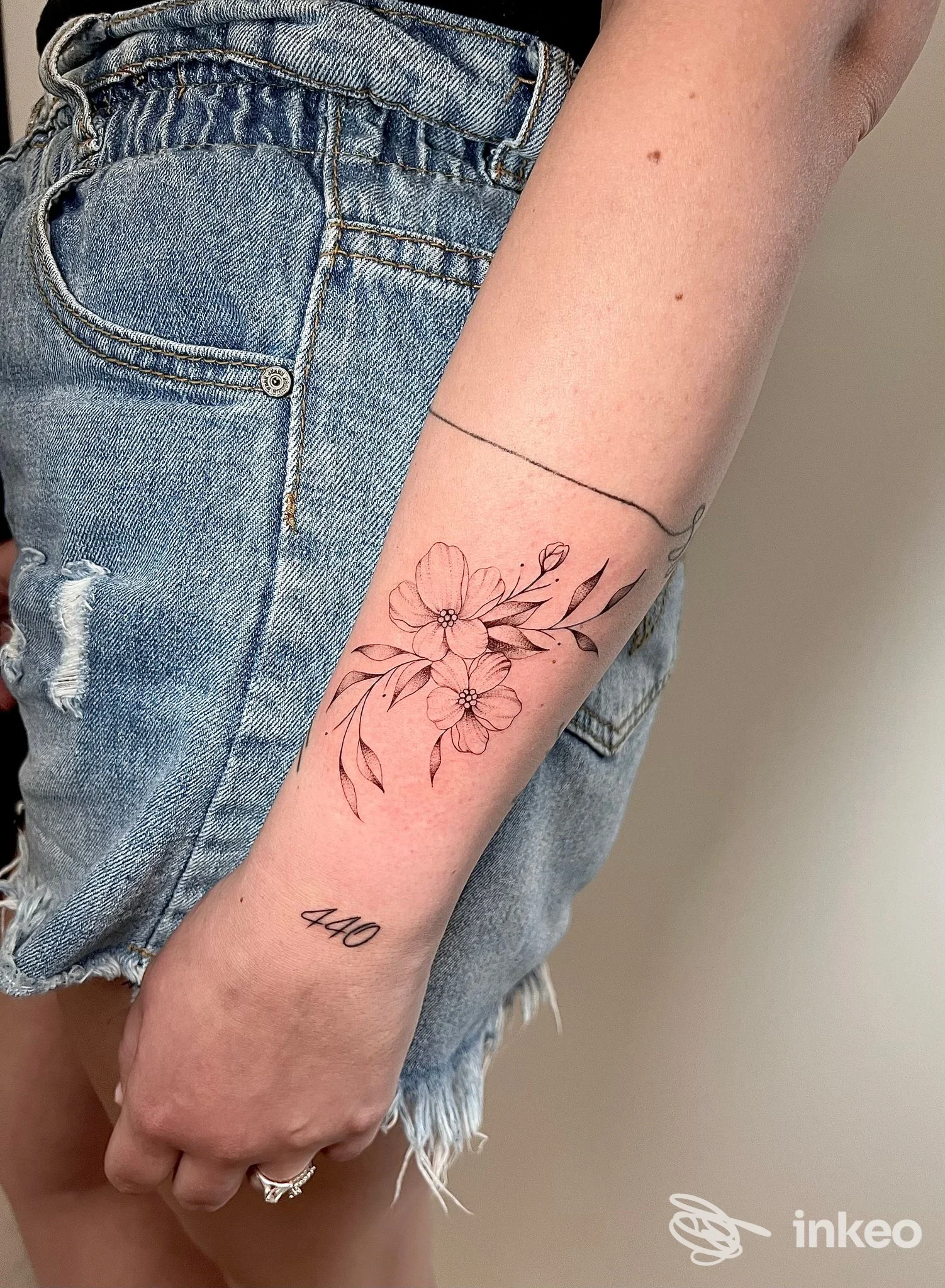 Primo piano dell'avambraccio con due tattoo: delicato motivo floreale lineare sulla pelle e una piccola scritta 440 sul polso; sfondo neutro, look casual con maniche di jeans.