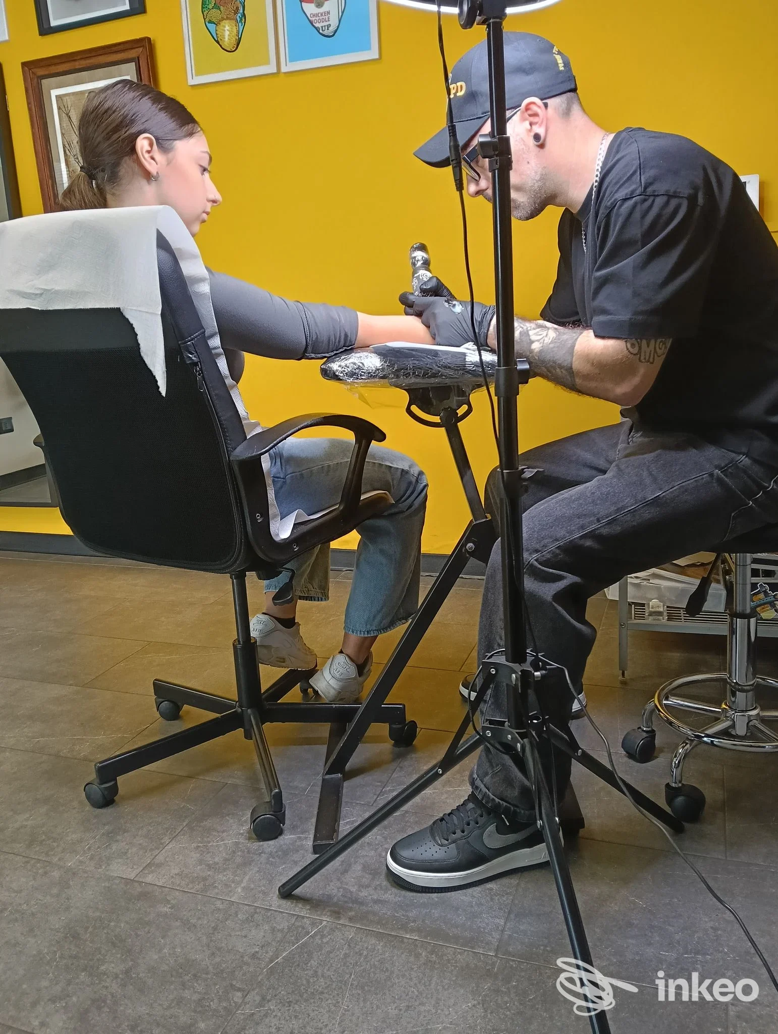 Un tatuatore con cappellino esegue un tatuaggio sulla mano di una cliente seduta in un salone con pareti gialle e cornici appese.