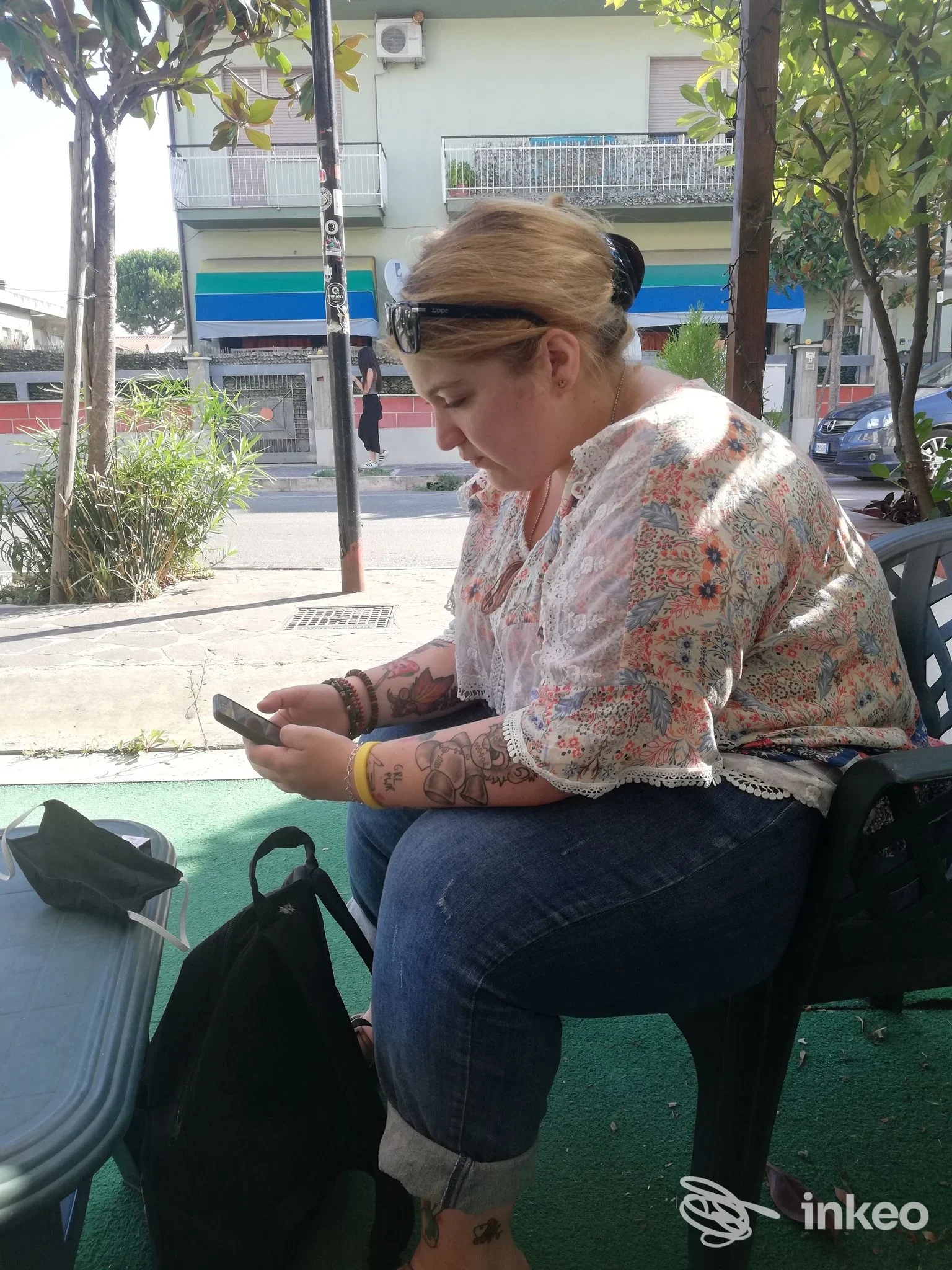 Donna seduta all'aperto su una sedia di plastica, guarda il telefono; è visibile un tatuaggio sulle braccia, sfondo urbano con tessuti e palazzi, luce di giorno; studio Italian Style Tattoo, Montesilvano.
