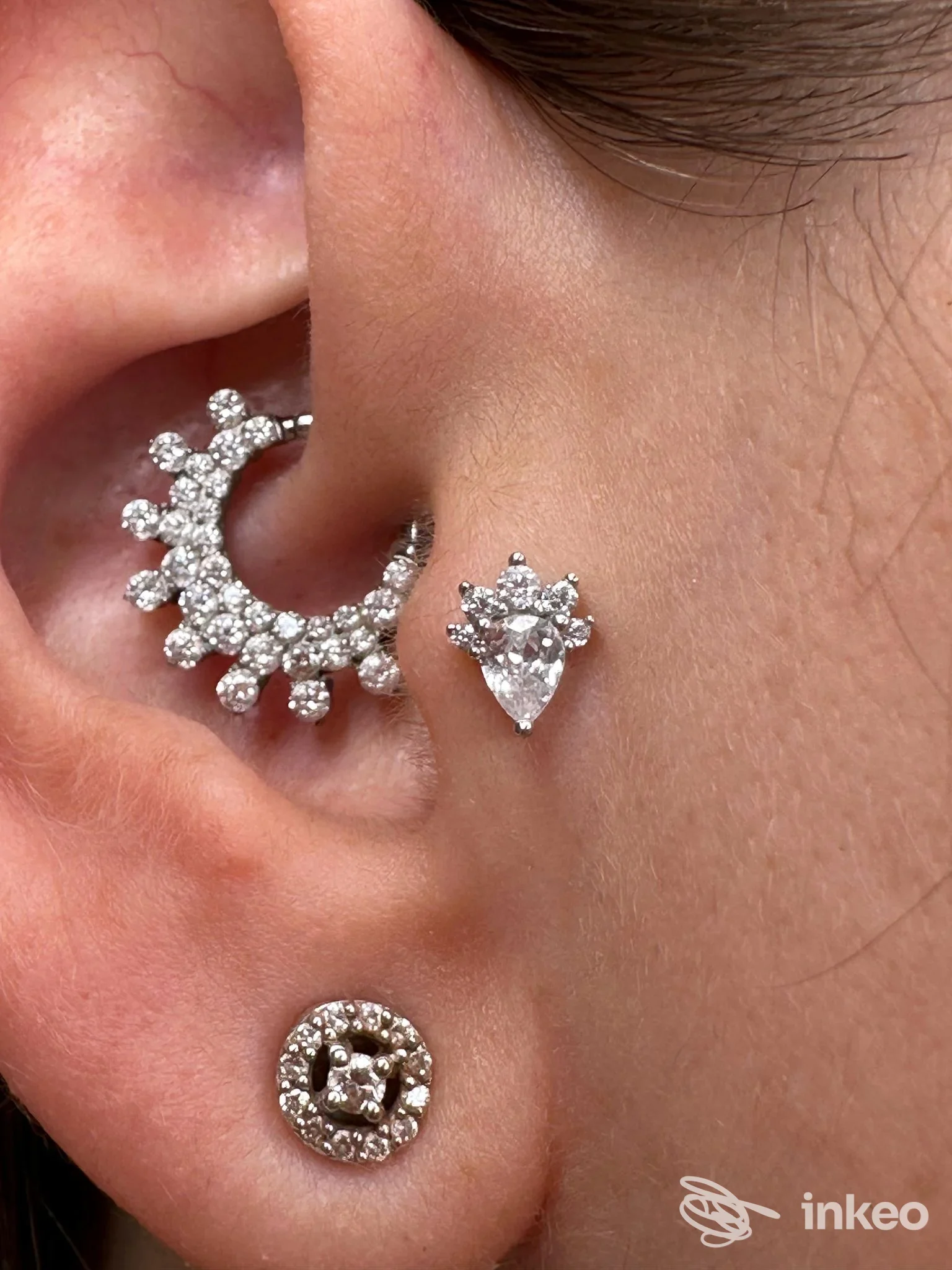 Orecchio sinistro con tre piercing: un cerchio ornato di zirconi sul bordo esterno, un piercing a forma di triangolo a lato e un piccolo foro dorato con zirconi sul lobo,