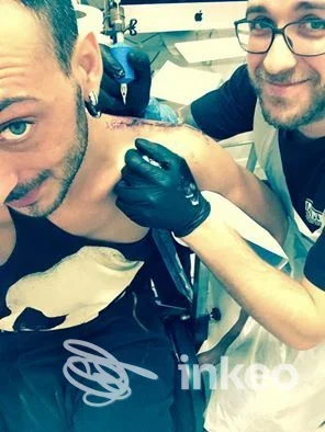 Due tatuaggio in corso sulla spalla di un cliente, mentre l'artista esegue l'iniezione sulla pelle; viso parziale dell'accompagnatore visibile, senza volto intero, senza watermark o testo di piattaforma.