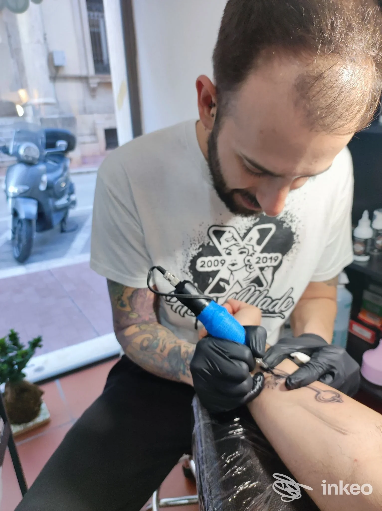 Un tatuaggio in corso su un braccio, realizzato nello studio Hand Made Tattoo Shop di Sambuceto; l’operatore indossa guanti neri e lavora con una macchina da tatuaggio, sfondo neutro e atmosfera di studio.