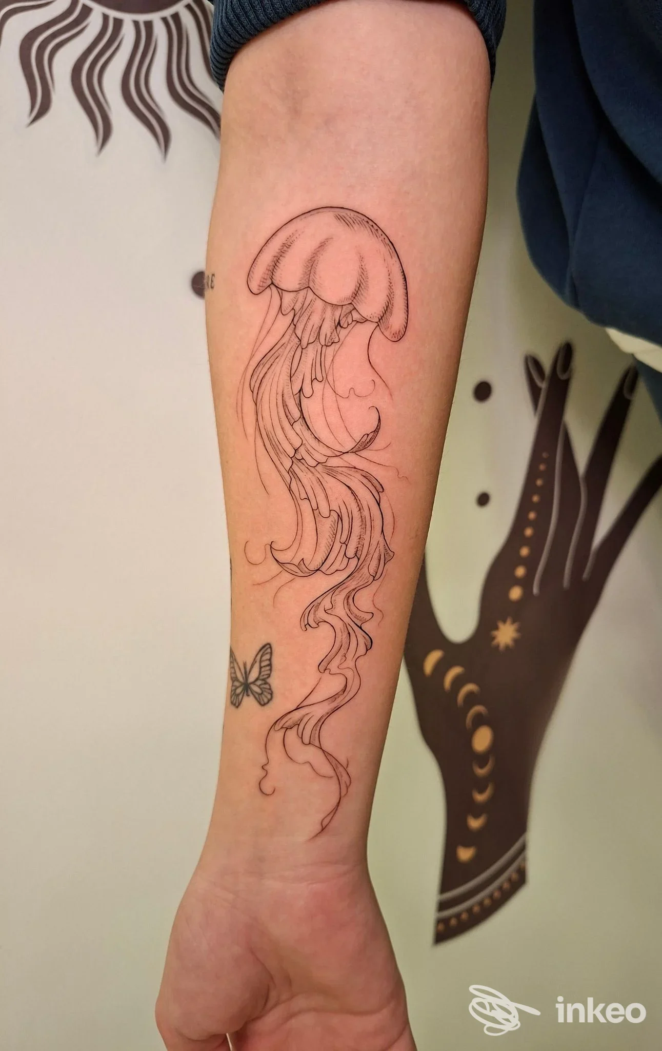 Avambraccio di una persona con tatuaggio a linee rosse rappresentante una medusa stilizzata che contiene linee ondulate, con sfondo chiaro; sullo sfondo si intravedono elementi decorativi.